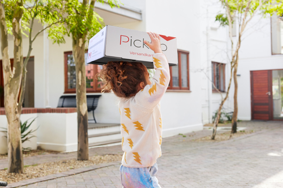 Mädchen trägt einen Pickup Versandkarton auf ihrem Kopf den Gehsteig entlang
