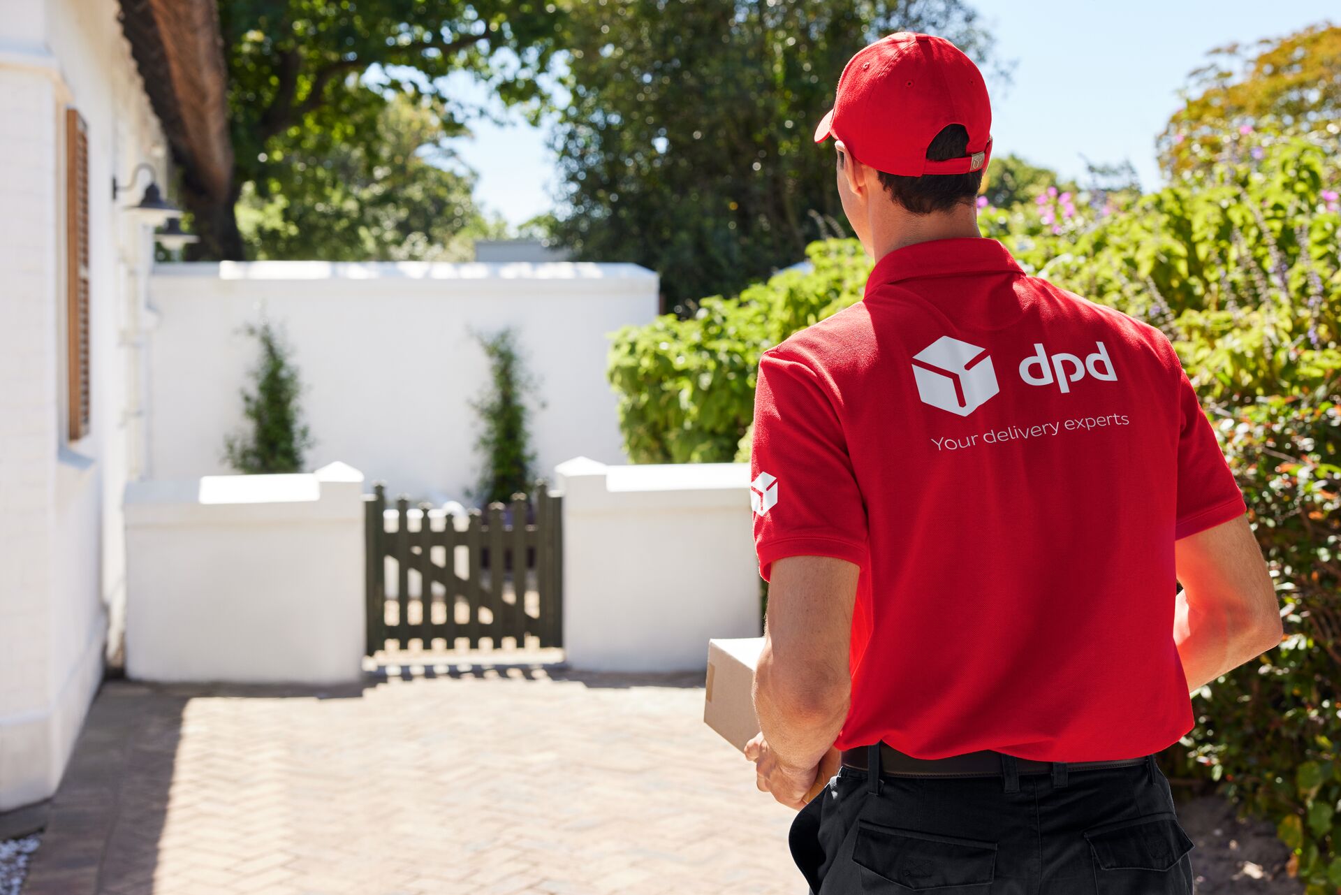 Kurier DPD z paczką idzie w stronę domu. Widoczne logo DPD na koszulce.