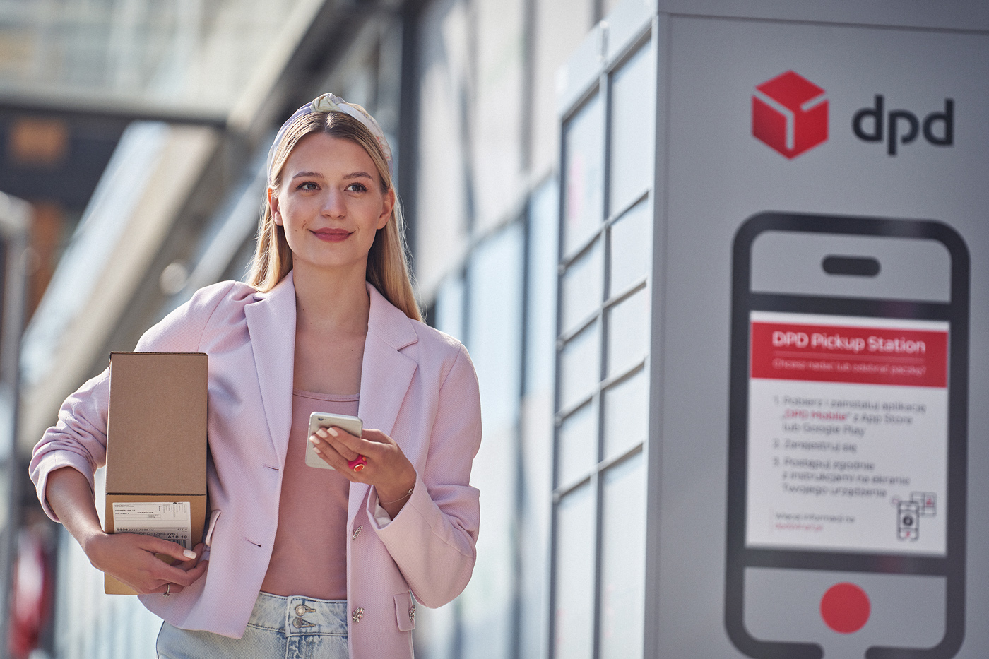 Kobieta z paczką i telefonem przy automacie paczkowym DPD. Odbiór przesyłki.