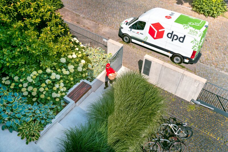 Kurier DPD z paczką idzie do klienta. W tle elektryczny samochód dostawczy DPD.