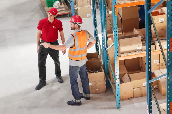 DPD News tresc 600×400 Pracownicy DPD w magazynie, jeden w uniformie, drugi w kamizelce i kasku.