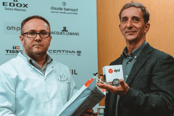DPD News tresc 600×400 duet Dwóch mężczyzn prezentuje model pojazdu DPD. Nagrody i logo marek w tle.