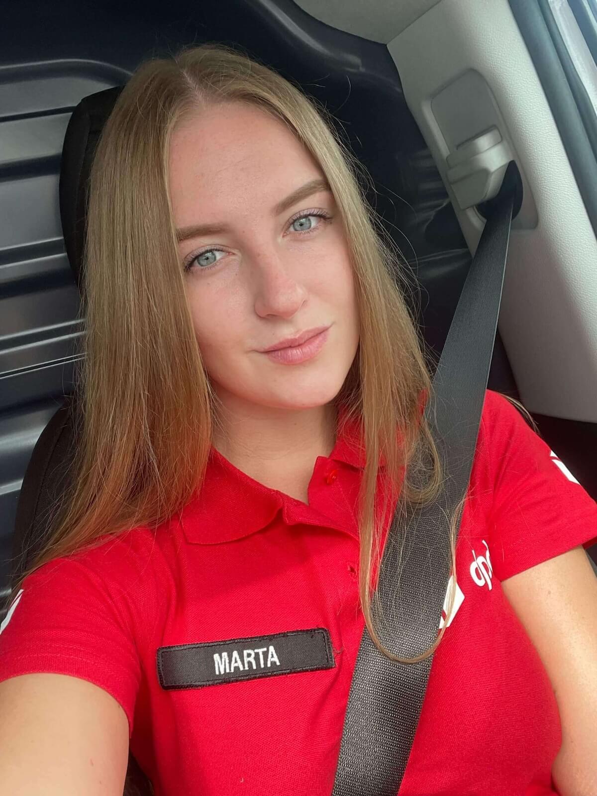 Kurierka DPD Marta w czerwonej koszulce z logo firmy, zapięta pasami w samochodzie.