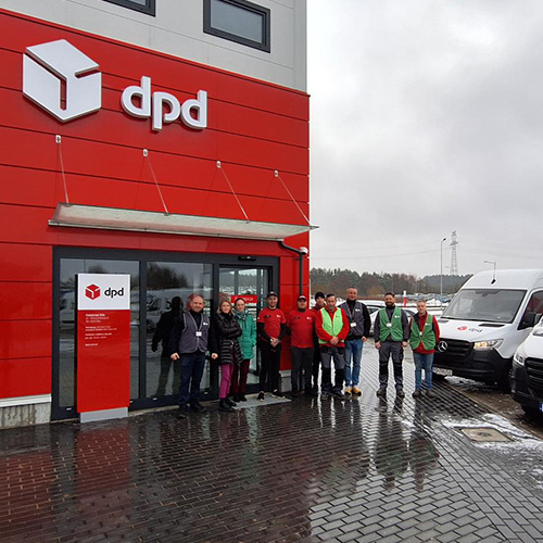 500×500 Grupa pracowników DPD przed budynkiem firmy i samochodami dostawczymi.
