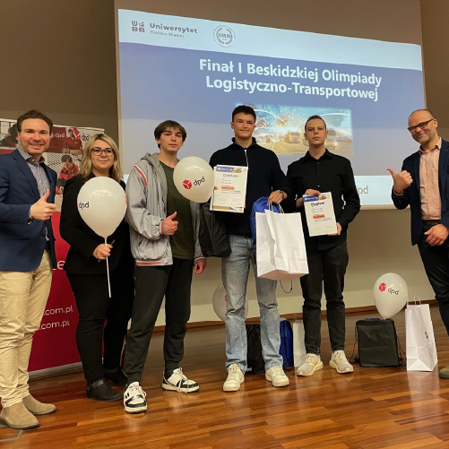 Laureaci I Beskidzkiej Olimpiady Logistyczno-Transportowej na scenie z organizatorami, trzymając dyplomy i nagrody, podczas ceremonii rozdania nagród na Uniwersytecie Bielsko-Bialskim.
