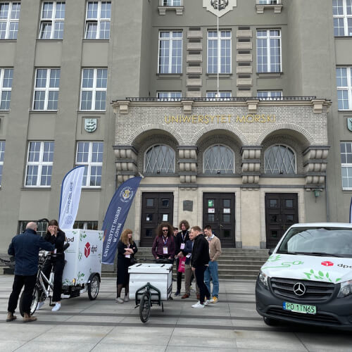 Grupa osób stojąca przed budynkiem Uniwersytetu Morskiego, obok samochodów i rowerów z logotypami DPD.