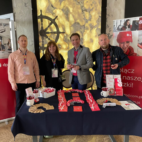 Pracownicy DPD przy stoisku promocyjnym podczas konferencji, z gadżetami i materiałami informacyjnymi na stole.