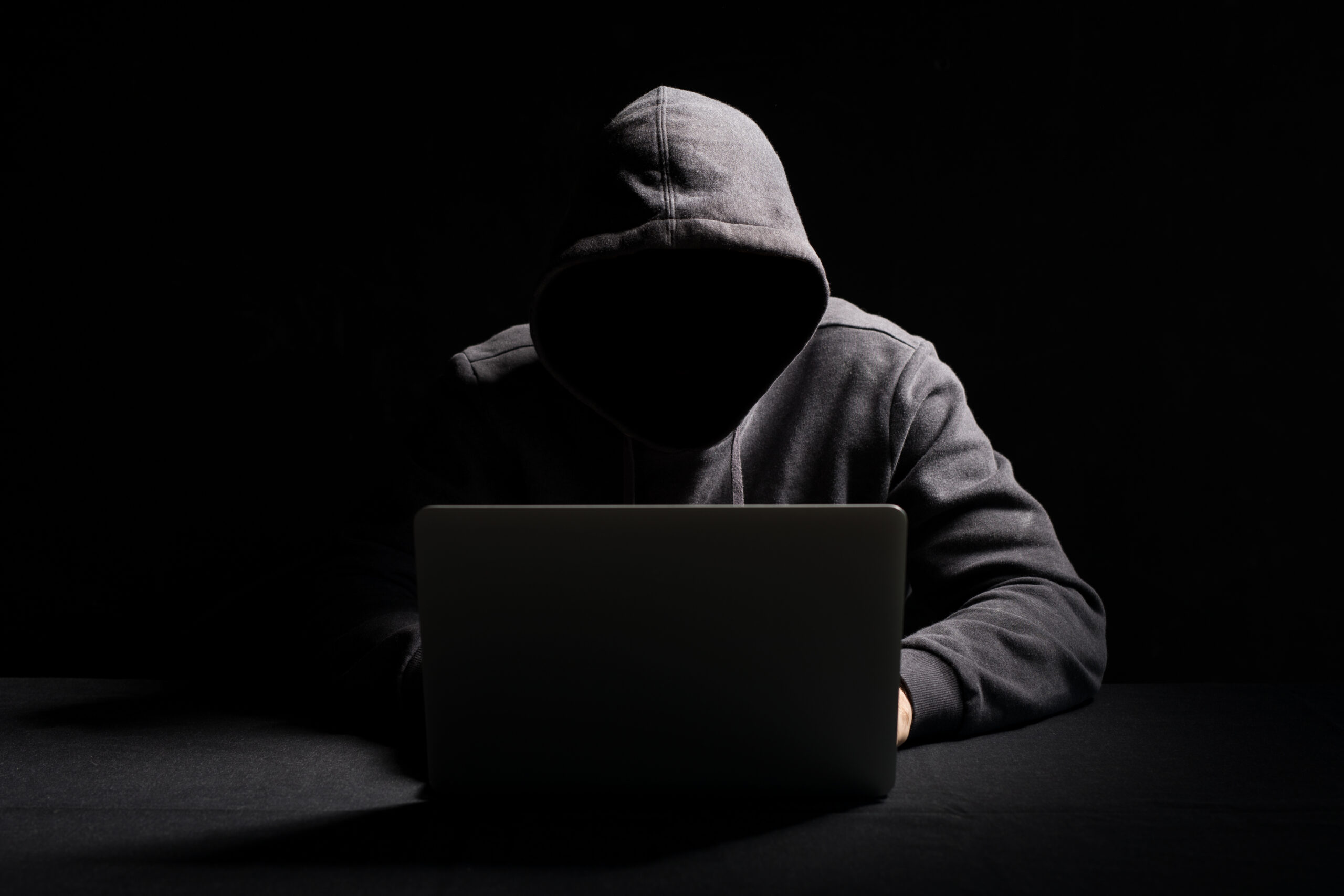poradnik_phishing_2 Haker w kapturze siedzący przed laptopem: Haker ubrany w ciemny kaptur pracujący przy laptopie w ciemnym otoczeniu.