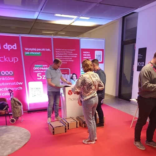 Stoisko DPD na targach Vivelo 2024, gdzie odwiedzający rozmawiają z pracownikami firmy. W tle widoczny jest baner promujący różne usługi DPD, a na stole leżą paczki i ulotki informacyjne.