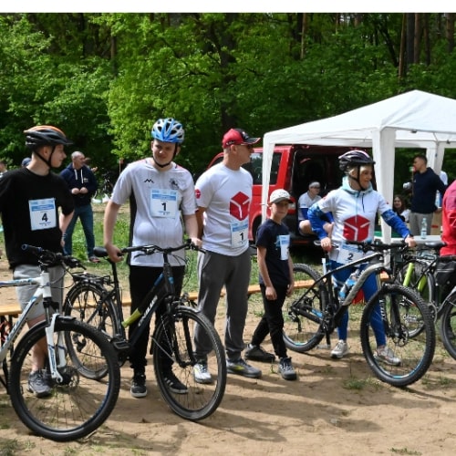 Grupa uczestników triathlonu młodzieżowego organizowanego przez DPD, stojąca obok rowerów i namiotu. Wszyscy mają na sobie kaski rowerowe i koszulki z logo DPD.