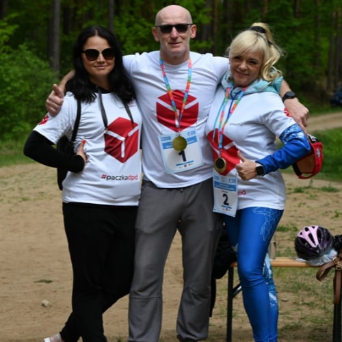Trzy osoby w koszulkach z logo DPD, uśmiechające się i obejmujące się, stojące na trasie triathlonu młodzieżowego. W tle widać las.