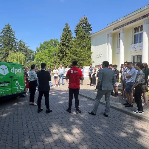 Projekt-bez-nazwy-31 (1) Grupa osób słucha prezentacji DPD przy samochodzie elektrycznym.