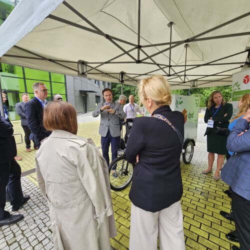 Spotkanie na świeżym powietrzu podczas konferencji Green Cities, gdzie przedstawiciel DPD Polska prezentuje rower elektryczny do dostaw. Grupa ludzi słucha prezentacji pod namiotem.