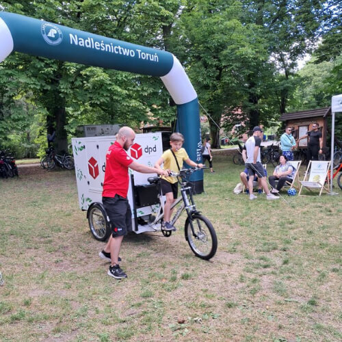 Pracownik DPD Polska prezentuje ekologiczny rower dostawczy podczas wydarzenia "Toru na Rowery 2024". Dziecko siedzi na rowerze, a w tle widać bramę startową Nadleśnictwa Toruń oraz innych uczestników wydarzenia.