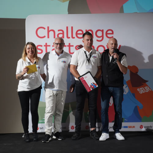 Jakub Biarda, wyróżniony jako najlepszy kurier w Grupie GeoPost, stoi na scenie wraz z innymi laureatami i organizatorami konkursu Challenge Starts Now. Na tle logotypu wydarzenia prezentują otrzymane nagrody i gratulacje.