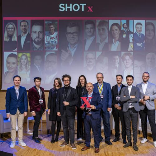 Grupa prelegentów i organizatorów SHOT x pozuje na scenie podczas festiwalu nauki e-x-plory w Gdyni. W tle widnieją portrety zaproszonych mówców.