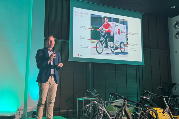 Maksymilian Pawłowski, kierownik ds. Komunikacji Korporacyjnej w DPD Polska, prezentuje rower cargo DPD na scenie podczas 5. edycji Polish Bicycle Summit. W tle widoczny jest slajd z opisem roweru i zdjęciem kuriera DPD na rowerze cargo. Na pierwszym planie widać stojące rowery elektryczne.
