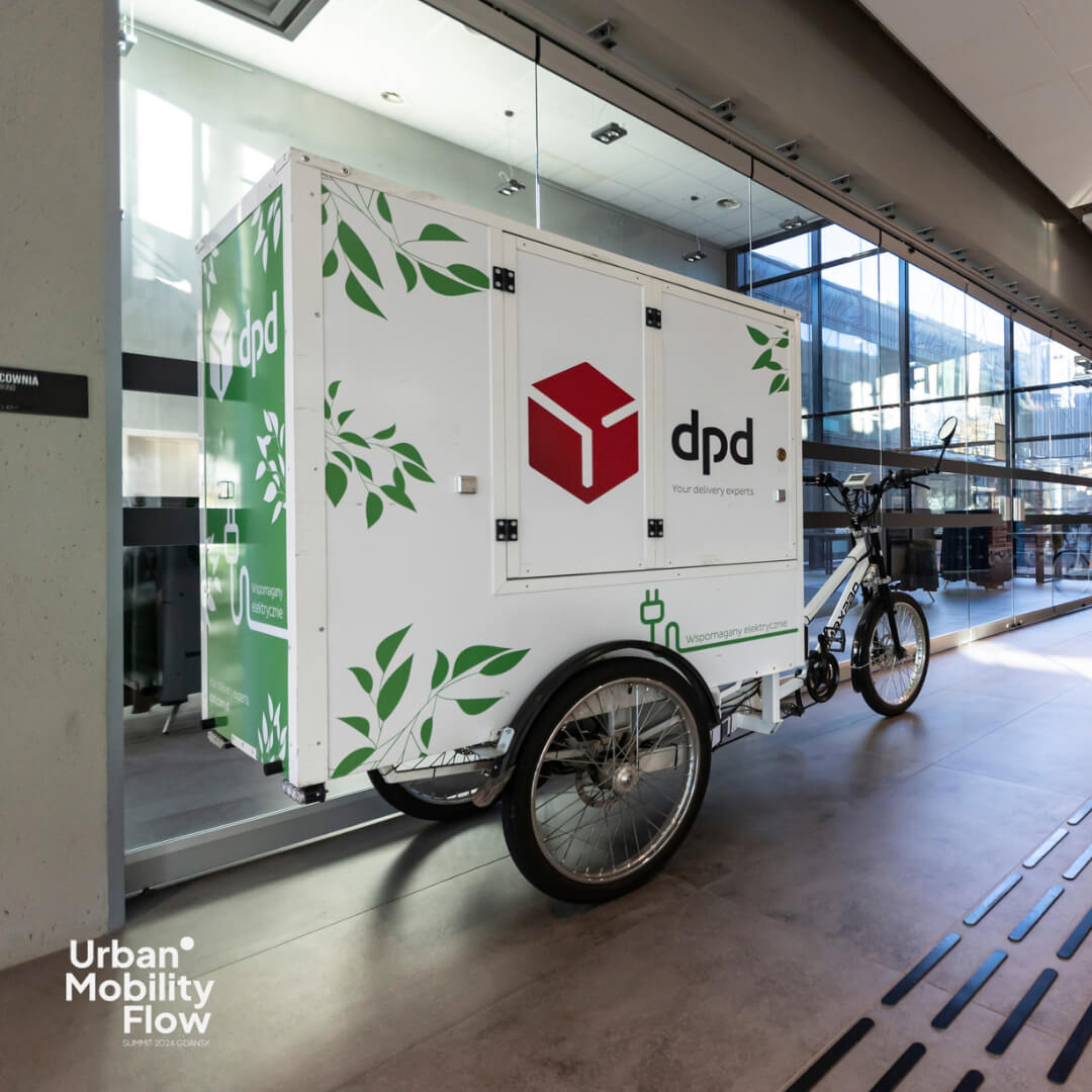 Projekt bez nazwy (77) Rower cargo należący do floty DPD Polska, ustawiony w przestrzeni konferencyjnej Urban Mobility Flow Summit. Pojazd oznaczony logotypem DPD i zielonymi wzorami, podkreślającymi ekologiczne podejście firmy.