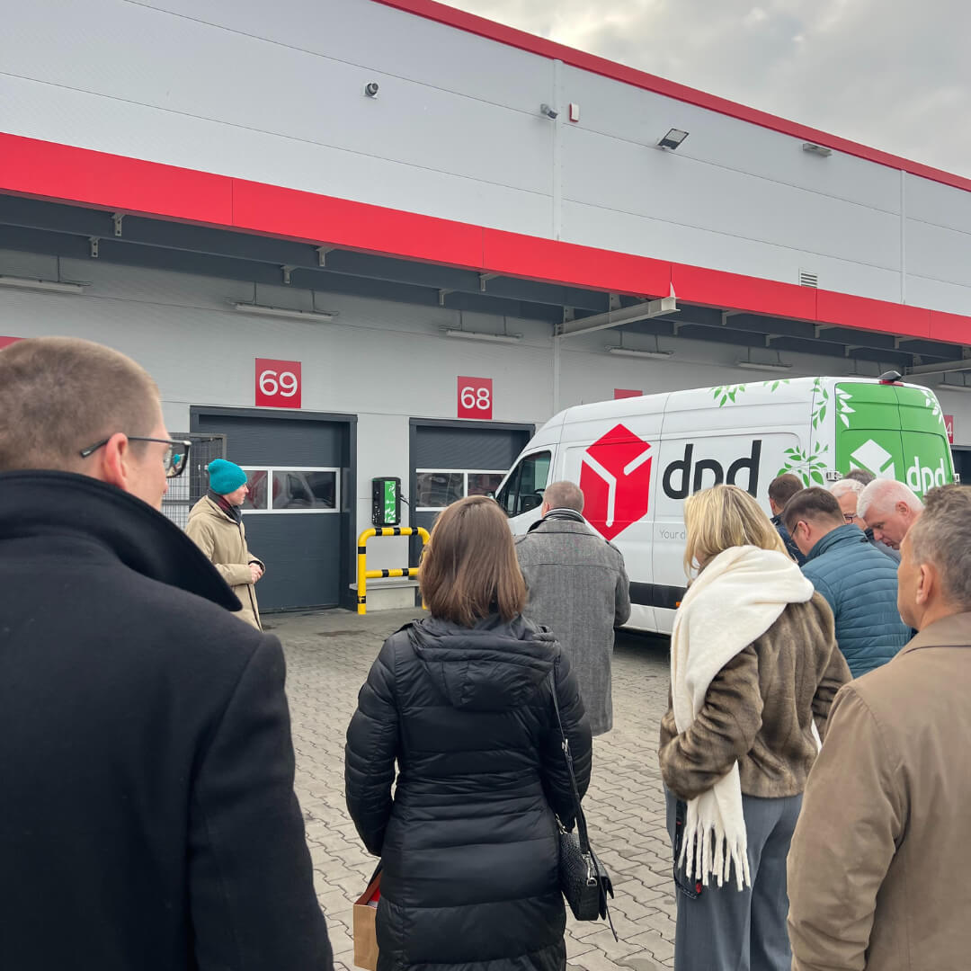 Grupa uczestników konferencji ogląda flotę elektrycznych samochodów dostawczych DPD zaparkowaną przy bramach oznaczonych numerami 68 i 69 w centrum logistycznym.