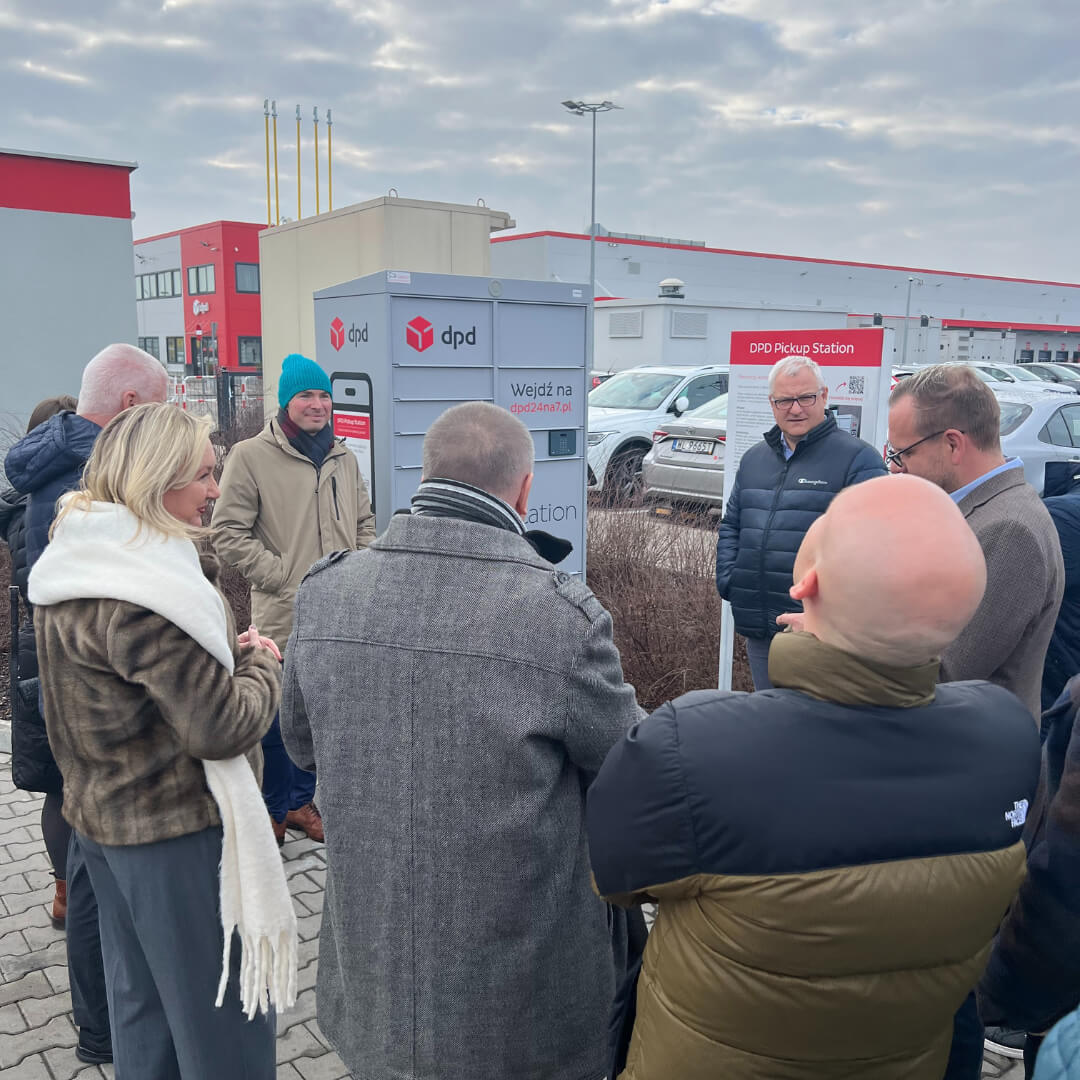Grupa osób ogląda nowoczesny automat paczkowy DPD Pickup Station, znajdujący się na zewnątrz budynku logistycznego. Prezentacja prowadzona jest przez pracownika firmy.