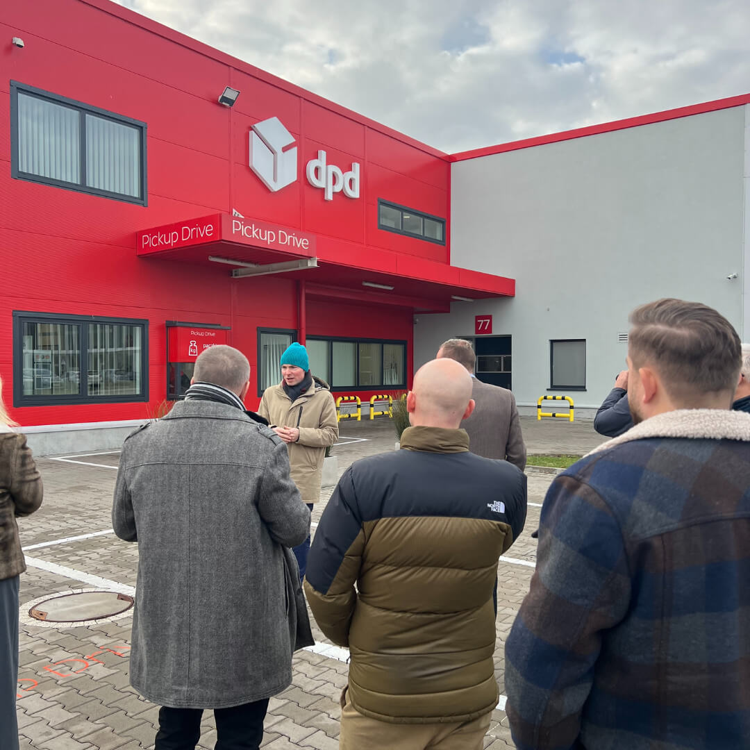 Grupa osób zebrana przed punktem DPD Pickup Drive, który umożliwia odbiór i nadawanie przesyłek bez konieczności opuszczania samochodu. Budynek z czerwonym logo DPD i dużymi oknami w tle.