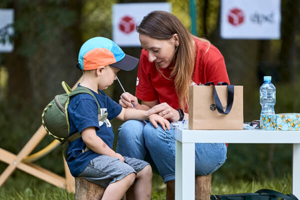 Wolontariuszka w czerwonej koszulce DPD maluje twarz chłopca w czapce z daszkiem i plecakiem, siedzącego na drewnianym pieńku. W tle widoczne są banery z logo DPD.