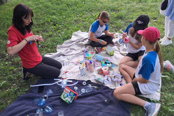 Kobieta w czerwonej koszulce DPD prowadzi warsztaty z dziećmi, które siedzą na kocu na trawie, wykonując zabawki z materiałów recyklingowych, takich jak plastikowe butelki i różnorodne ozdoby.