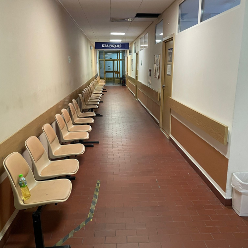 Projekt bez nazwy (99) Korytarz szpitalny z rzędem krzeseł i napisem "Izba Przyjęć" w oddali.