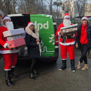 Kurierzy DPD w strojach Świętego Mikołaja z prezentami przy elektrycznym samochodzie.