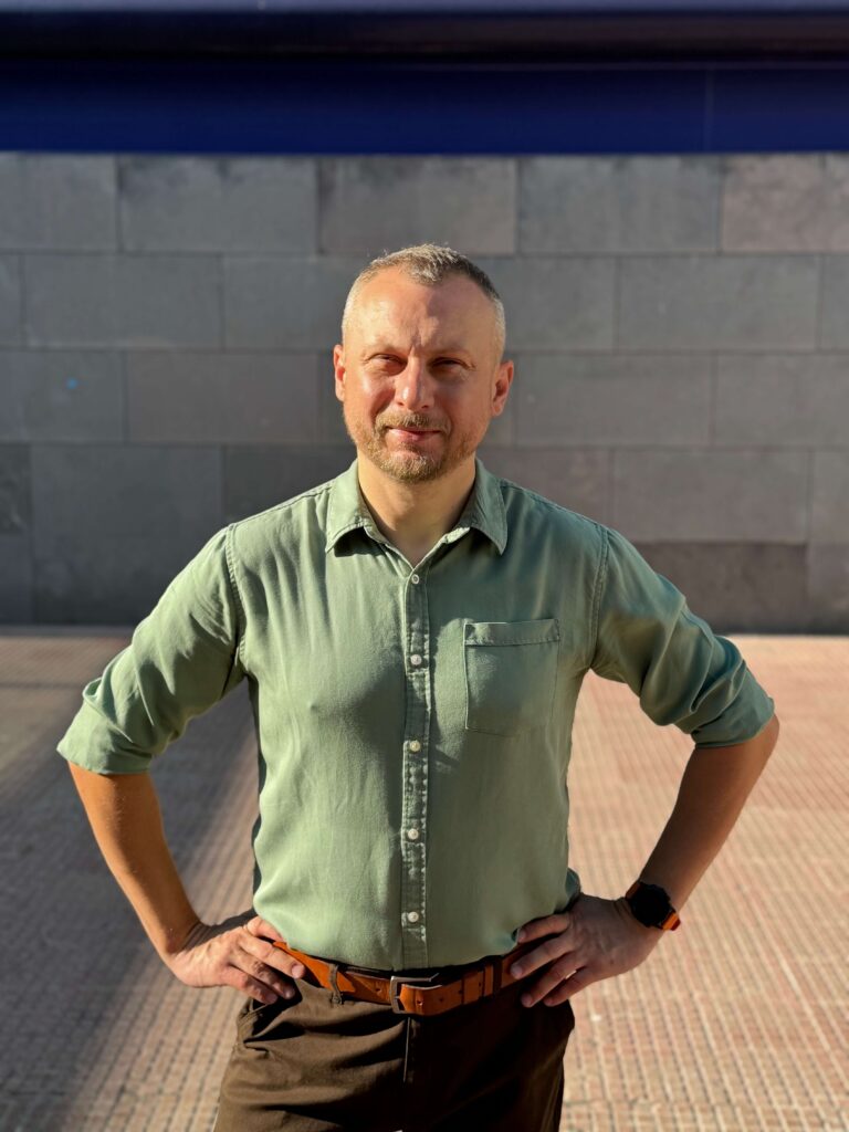 Mężczyzna w zielonej koszuli i brązowych spodniach z założonymi rękami na tle murku