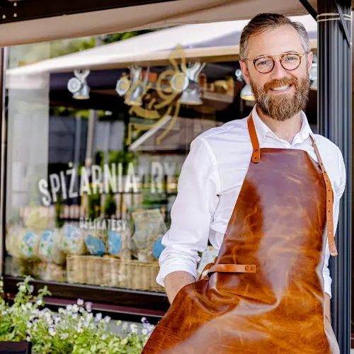 Michał Błaszczyk, założyciel Spiżarni Rydzyńskiej, w skórzanym fartuchu przed swoim sklepem z ekologiczną żywnością. W tle witryna z napisem Spiżarnia Rydzyńska Delikatesy.