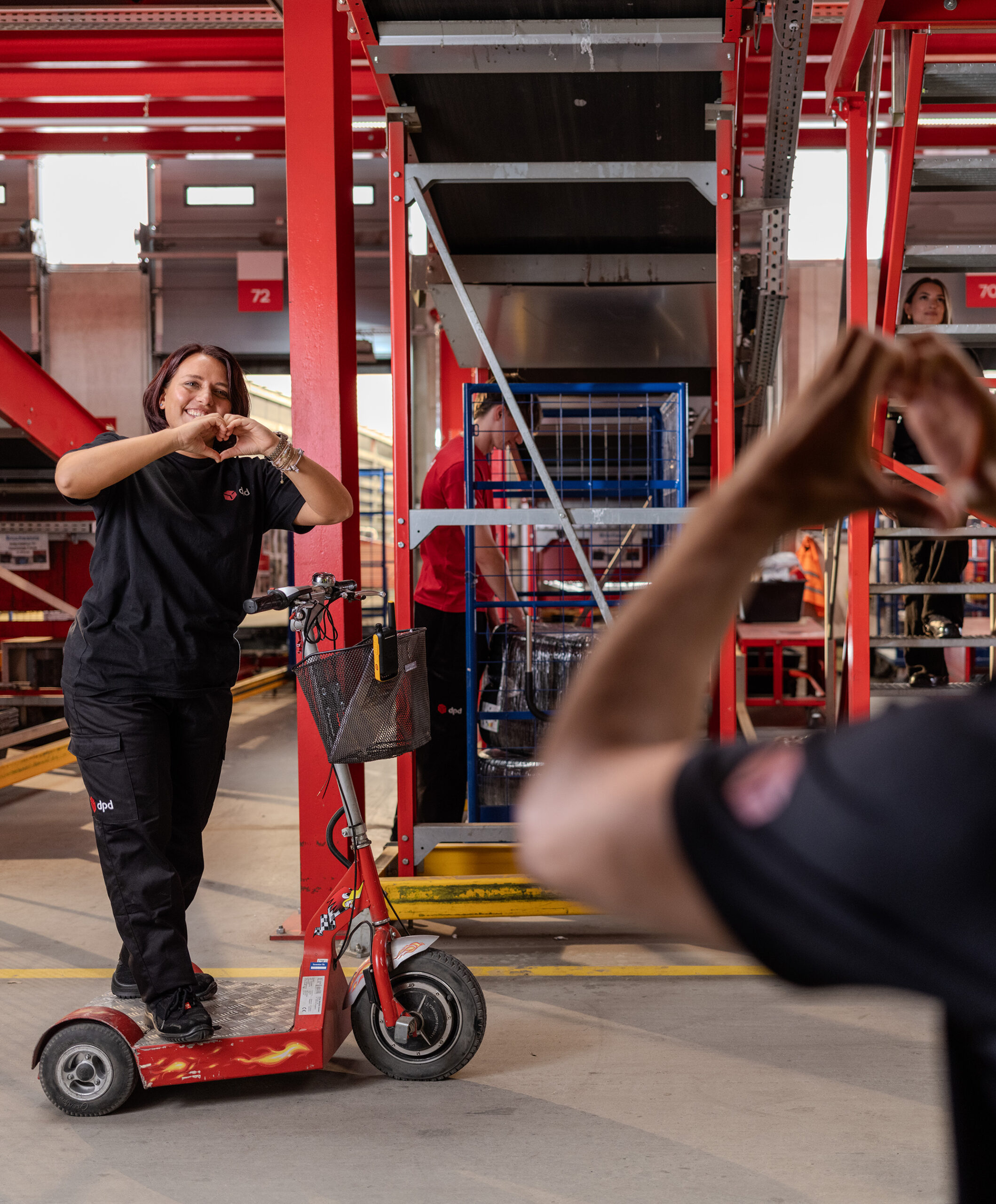 Frau fährt auf einem Scooter durch das Depot und begrüßt herzlich einen Kollegen.