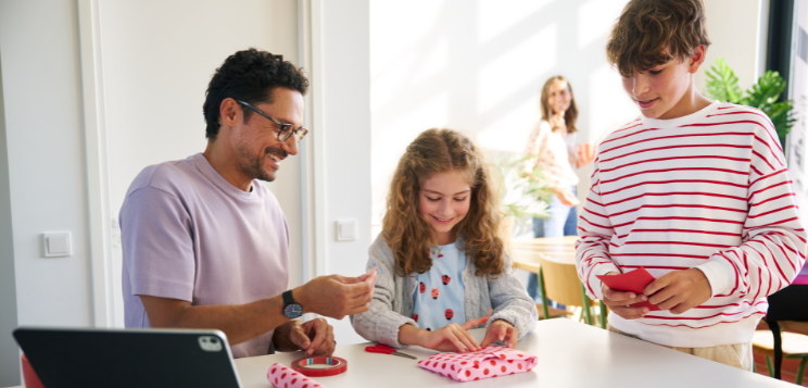 Ein Vater sitzt mit seinen Kindern am Tisch und packt lachend Geschenke in buntes Papier ein