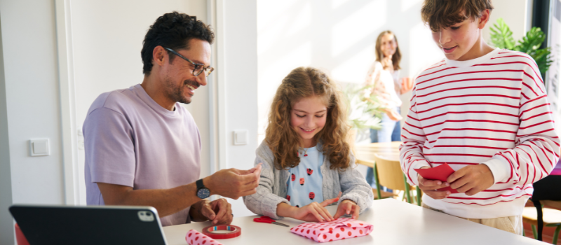 Ein Vater sitzt mit seinen Kindern am Tisch und packt lachend Geschenke in buntes Papier ein
